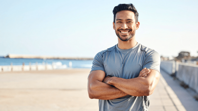 A man with a warm smile and short dark hair stands outdoors with his arms crossed. He is wearing a grey athletic t-shirt, and a coastal walkway with a blue ocean and clear sky is visible in the background.