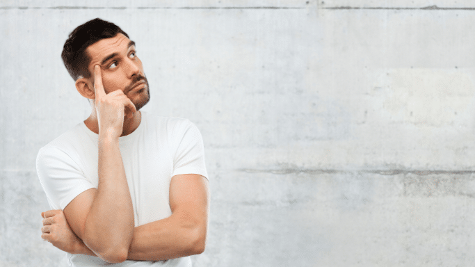 A man in a white t-shirt looking upward and to the side with a thoughtful expression. He has one hand raised to his temple as if in deep concentration, posed against a textured light grey concrete wall.