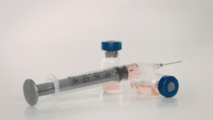 A close-up of a medical syringe lying horizontally in front of two glass vials containing a light pink liquid. The background is a clean, neutral white.