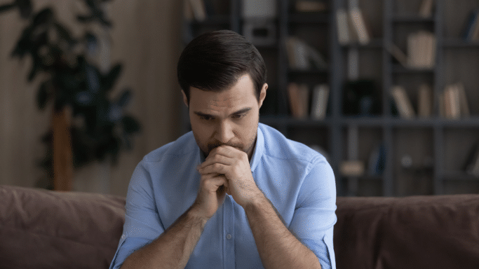 A medium shot of a man sitting on a sofa, looking pensive or concerned. He has his hands clasped in front of his face and is looking downward, suggesting worry or deep thought.