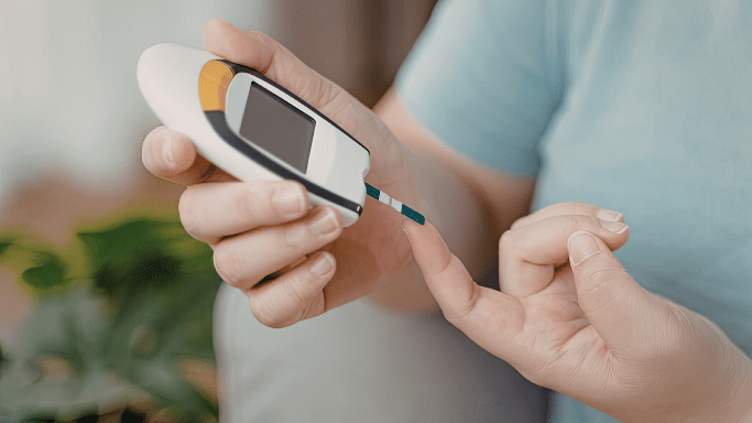 A close-up of a person’s hands using a glucose meter. They are holding the device and applying a drop of blood from their fingertip to a test strip to check their blood sugar levels.
