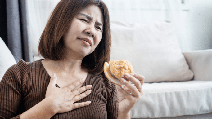 A woman sitting on a floor, holding a bitten burger. She has a pained or uncomfortable expression, with one hand pressed against her chest, suggesting she is experiencing heartburn or indigestion.