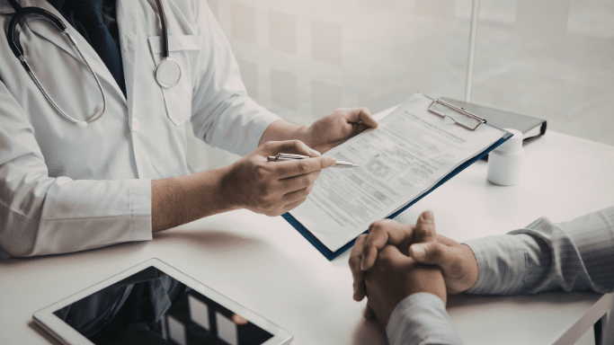 A professional setting showing a doctor in a white lab coat sitting at a desk. The doctor is pointing to a medical document on a clipboard while speaking with a patient, whose hands are clasped on the table.