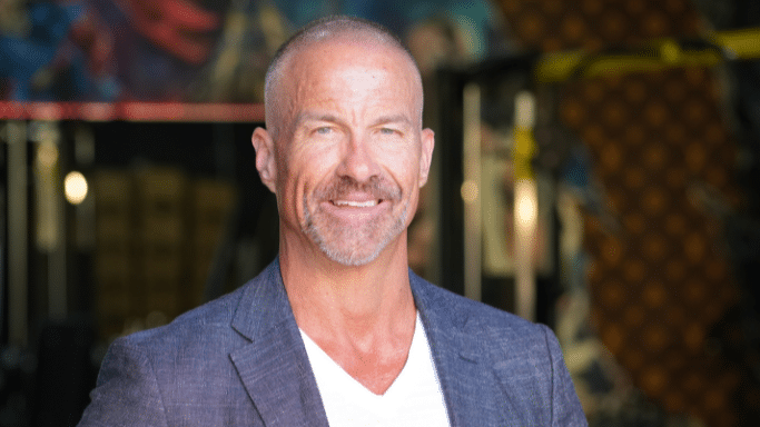 A portrait of a middle-aged man with a shaved head and a light beard. He is smiling confidently at the camera, wearing a white V-neck shirt and a grey blazer, representing a healthy, positive outcome.