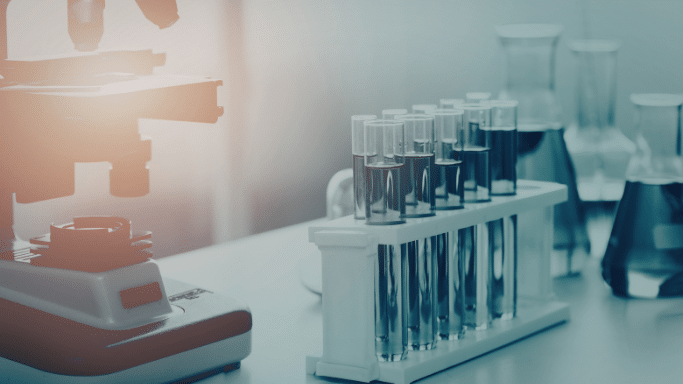 A wide-angle, atmospheric shot of a laboratory bench. On the left, the base and stage of a microscope are partially illuminated by a warm, orange flare. In the center, a white rack holds several glass test tubes filled with a dark blue liquid. The background is blurred, showing additional laboratory glassware like Erlenmeyer flasks, all bathed in a cool, teal-tinted light.