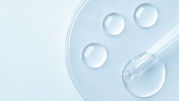 A minimalist, top-down view of a clear petri dish resting on a light blue surface. Inside the dish are three perfect, transparent liquid droplets. A glass pipette enters from the right, with its tip resting in a larger pool of clear liquid, creating a clean, clinical aesthetic.