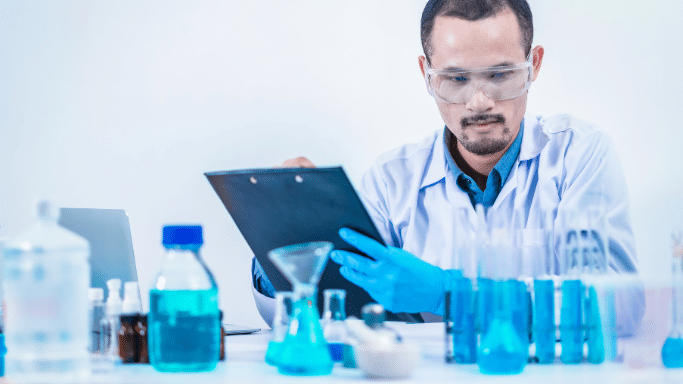 A male scientist with a goatee, wearing a white lab coat, blue shirt, safety goggles, and blue nitrile gloves. He is focused on a clipboard he holds in his hands. In the foreground, the lab bench is crowded with glassware containing vibrant blue and teal liquids, including beakers, flasks, and test tubes.