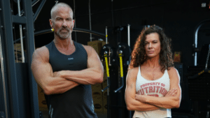 Man and woman standng in the gym with their arms crossed, looking directly at the camera with serious expressions.