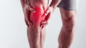 A close-up of a person's legs while they are standing. Both hands are gripping their right knee, which is highlighted with a bright red glowing effect to symbolize intense pain or inflammation.