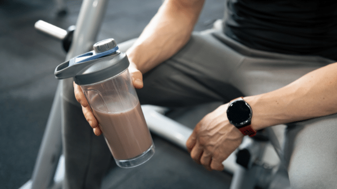A close-up view of a person in a gym holding a transparent shaker bottle filled with a chocolate-colored protein shake. They are wearing a red and black smartwatch.