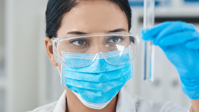 A close-up shot of a female scientist wearing a white lab coat, a blue surgical mask, and clear safety goggles. Her eyes are focused intently on a glass test tube filled with clear liquid that she is holding up with a gloved hand. The lighting is bright and professional.