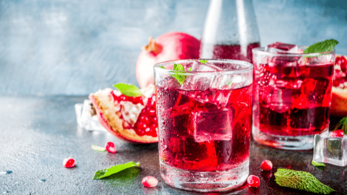 Two glasses of deep red pomegranate juice garnished with fresh mint leaves and ice cubes. The glasses are on a dark, textured surface next to a whole pomegranate and a piece of sliced pomegranate.
