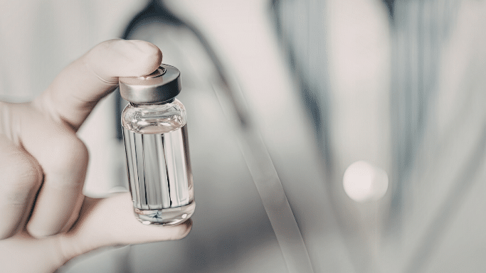 A close-up, slightly blurred shot of a hand in a white surgical glove holding a small glass medical vial with a silver cap. The vial contains a clear liquid. The background is out of focus, showing the white coat and stethoscope of a medical professional, emphasizing the theme of healthcare or vaccination.