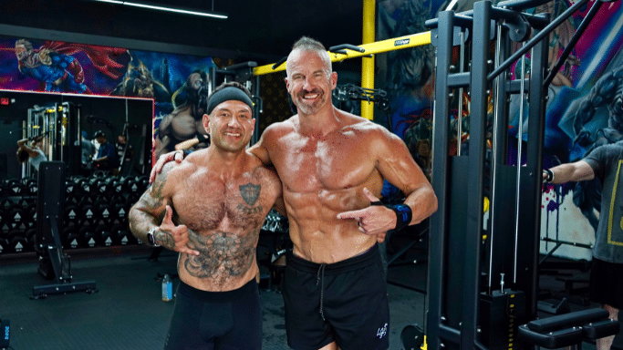 Two shirtless bodybuilders pose together in a gym. Both are smiling and gesturing toward each other. The gym background features colorful murals of comic book characters.