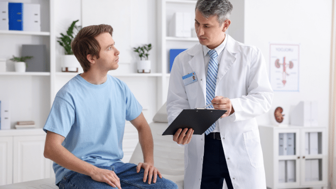 A male doctor in a white lab coat and blue tie speaks to a young male patient sitting on an examination table. The doctor holds a black clipboard and a pen.