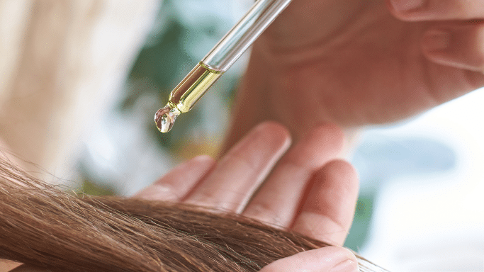 A close-up, high-angle shot shows a glass dropper releasing a single, golden drop of hair oil onto a section of brown hair held in a person's hand. The background is softly blurred.