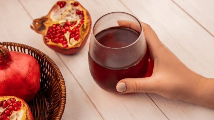 A close-up, high-angle shot of a hand holding a glass of dark red juice. Fresh pomegranates, both whole and halved, are visible on a white wooden table nearby.