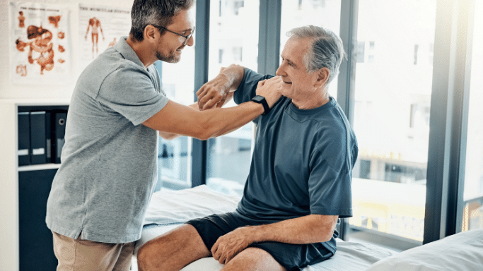 A physical therapist in a grey shirt and glasses works with an elderly man sitting on an examination table. The therapist is gently guiding the man's arm, and both are smiling, depicting a positive rehabilitation session.
