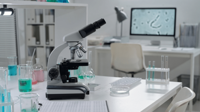 A clean, modern laboratory setting featuring a white microscope in the foreground. In the background, a computer monitor displays a microscopic image of a curved cell, surrounded by various glass beakers and test tubes filled with colorful liquids.