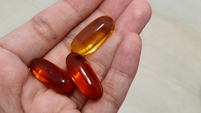 Three large, translucent liquid gel capsules—two amber-colored and one reddish-brown—resting in the palm of a person's hand.