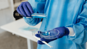 A close-up of a person in a blue laboratory gown and purple gloves. They are pouring a bright blue liquid from a graduated cylinder into a glass petri dish containing two small, white rectangular solids.
