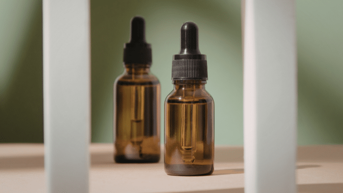 Two amber glass apothecary bottles with black dropper tops sit on a beige surface. The lighting creates soft shadows against a muted sage-green background, framed by two vertical white pillars.