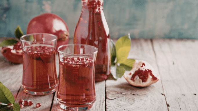 Two glasses of pomegranate juice filled with fresh pomegranate seeds, set on a rustic wooden table. A bottle of the juice and fresh pomegranate fruit decorate the background.