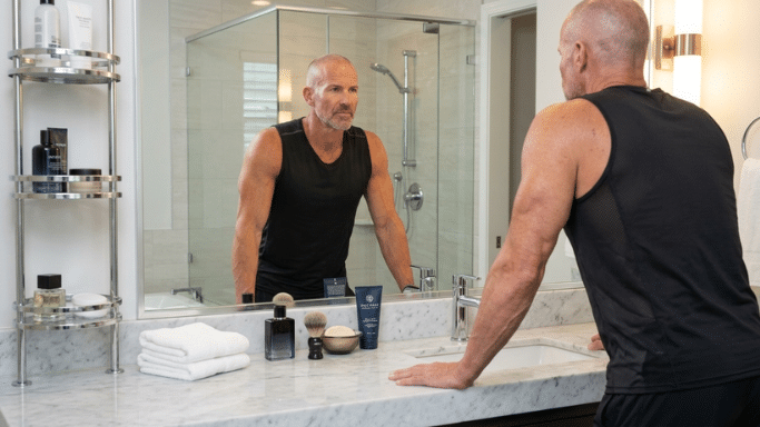A middle-aged man with a shaved head and an athletic build stands in a bright, modern marble bathroom. He is wearing a black sleeveless shirt and leaning against the counter, looking at his reflection in a large mirror.