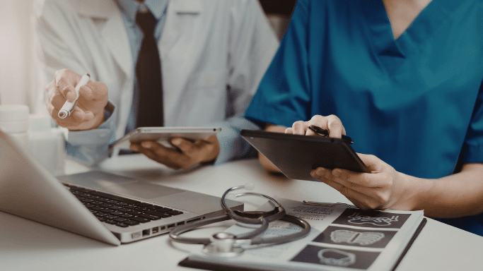 A close-up of two medical professionals working at a desk. One person in a white lab coat points with a pen at a laptop screen, while the other in blue scrubs uses a tablet.