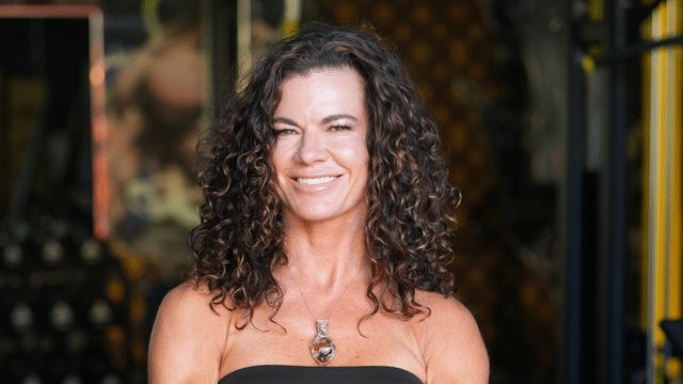 A portrait of a smiling woman with voluminous, dark curly hair and sun-kissed highlights. She is wearing a black strapless top and a teardrop-shaped pendant necklace, with a blurred dark background behind her.