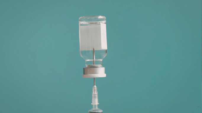 A clear medical vial sits inverted on a teal background, with a needle from a syringe inserted through the rubber stopper to draw out the liquid.