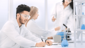 A bright, high-key photo of young scientists working in a modern lab. In the foreground, a man with a beard and safety goggles writes in a notebook. In the background, other researchers in white coats are blurred, one pointing toward a microscope.