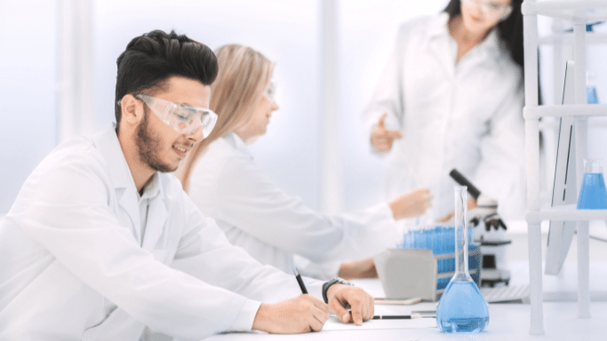 A bright, high-key photo of young scientists working in a modern lab. In the foreground, a man with a beard and safety goggles writes in a notebook. In the background, other researchers in white coats are blurred, one pointing toward a microscope.