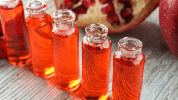A row of small glass vials filled with a bright orange-red liquid, possibly essence or oil, arranged diagonally on a light wooden surface. In the blurred background, a sliced open pomegranate with visible red seeds and a whole red apple are used as decorative accents.