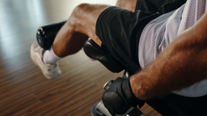 A close-up, low-angle shot of a person's legs using a leg extension machine in a gym. The focus is on the muscular quadriceps as they engage against the padded bar of the machine. The person is wearing black shorts, white socks, and light-colored sneakers.
