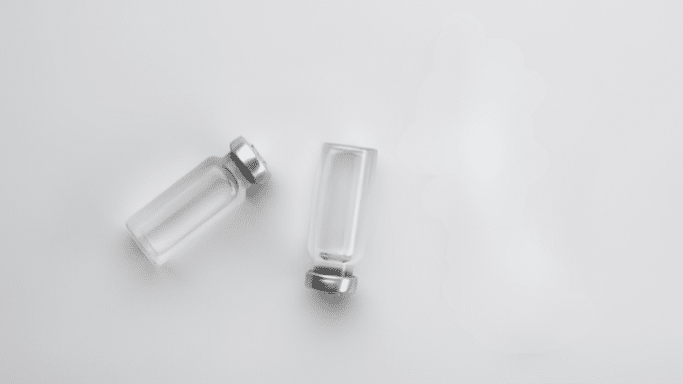 A minimalist top-down shot of two small, clear glass medical vials with silver caps resting on a plain white surface. A faint, translucent watery spill or shadow is visible to the right of the vials.