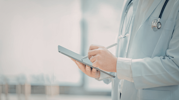 A mid-section view of a doctor in a white lab coat with a stethoscope around their neck. They are using a white stylus to write or navigate on a slim grey tablet.