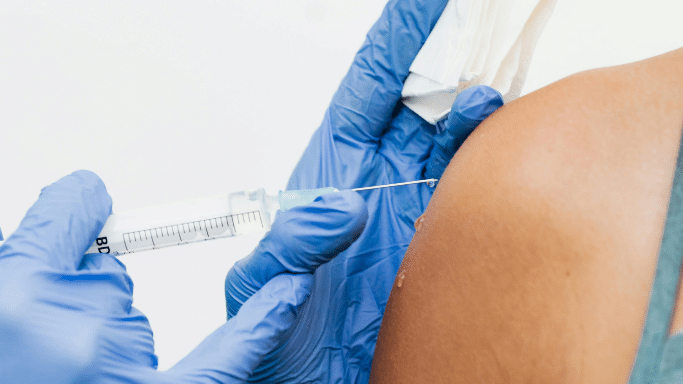 A close-up shot of a healthcare provider wearing blue nitrile gloves administering an injection into a person's upper arm. A small drop of liquid is visible on the needle tip, and a white cotton swab is held ready.