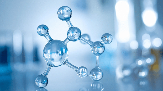 A clear, stylized molecular model made of glass-like spheres and connecting rods. The structure is set against a soft blue, out-of-focus laboratory background.