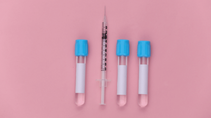 A flat-lay photograph on a pink background showing three medical test tubes with blue caps and white labels. A single clear syringe with a needle is placed vertically between the first and second tubes.