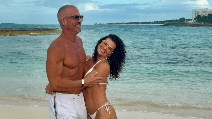 A candid shot of the couple on the same beach. They are embracing and laughing. The man is shirtless in white trunks and sunglasses, while the woman is in a white bikini and orange-tinted sunglasses.