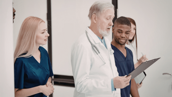 A senior doctor with a white beard and lab coat stands in a hallway, looking down at a clipboard. He is surrounded by three younger medical students or interns in blue and white scrubs who are listening attentively.