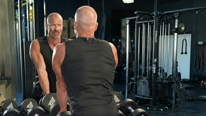 A muscular man with a shaved head seen from behind, looking at his reflection in a large gym mirror. He is resting his hands on a row of dumbbells, with various gym cables and weight machines visible in the background.