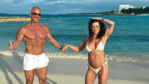 A man and woman with highly muscular, athletic physiques posing on a sunny beach. Both are smiling, wearing swimwear and sunglasses, and holding hands. A coastal resort is visible in the background under a blue sky.