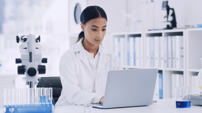 A female scientist in a white lab coat sits at a clean, modern lab bench, focused on her silver laptop. To her left is a microscope and a rack of blue-filled test tubes.