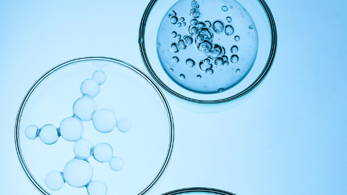 A top-down, artistic macro shot of three glass petri dishes on a light blue background. The dishes contain clear liquids with varying sizes of suspended air bubbles and oil-like droplets forming molecular patterns.