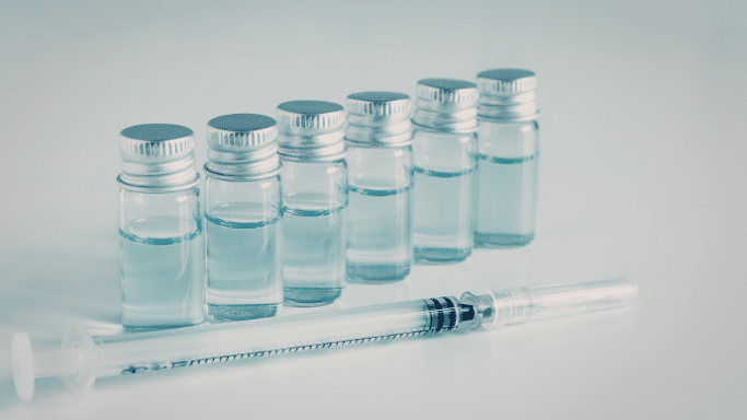 A clean, clinical shot of several small glass vials containing clear liquid, arranged in a row with a medical syringe laying in the foreground on a white surface.