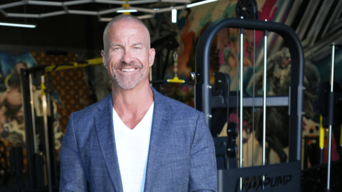A portrait of a middle-aged man with a short beard and graying hair, wearing a blue blazer and white t-shirt, smiling inside a gym setting.