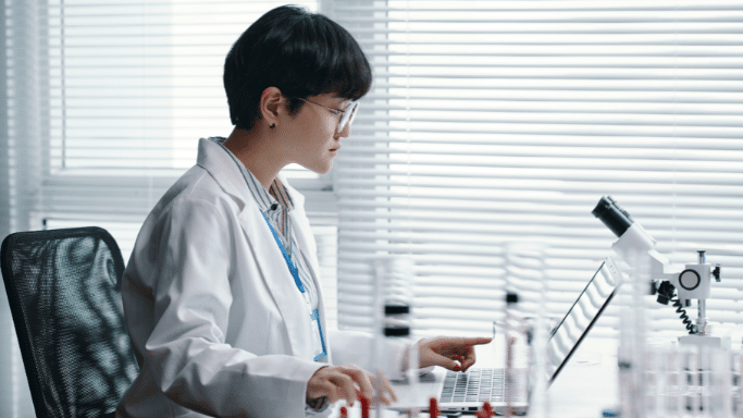 A scientist with short dark hair and glasses sits in a bright laboratory, working on a laptop. Various glass laboratory equipment and a microscope are visible on the desk against a backdrop of white window blinds.