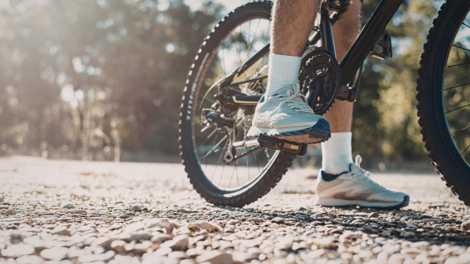 A low-angle, close-up shot of a person's legs while seated on a mountain bike. They are wearing white socks and light-colored sneakers, with one foot resting on a pedal. the background is a blurred, sunlit wooded area.